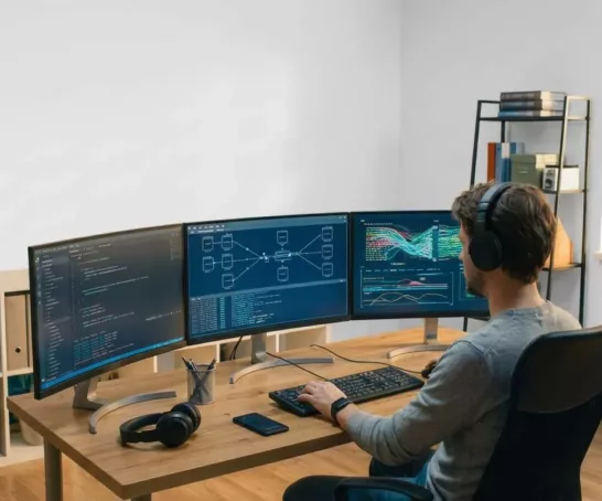 Entwickler mit Kopfhörern arbeitet an mehreren Monitoren mit Code an seinem Schreibtisch.
