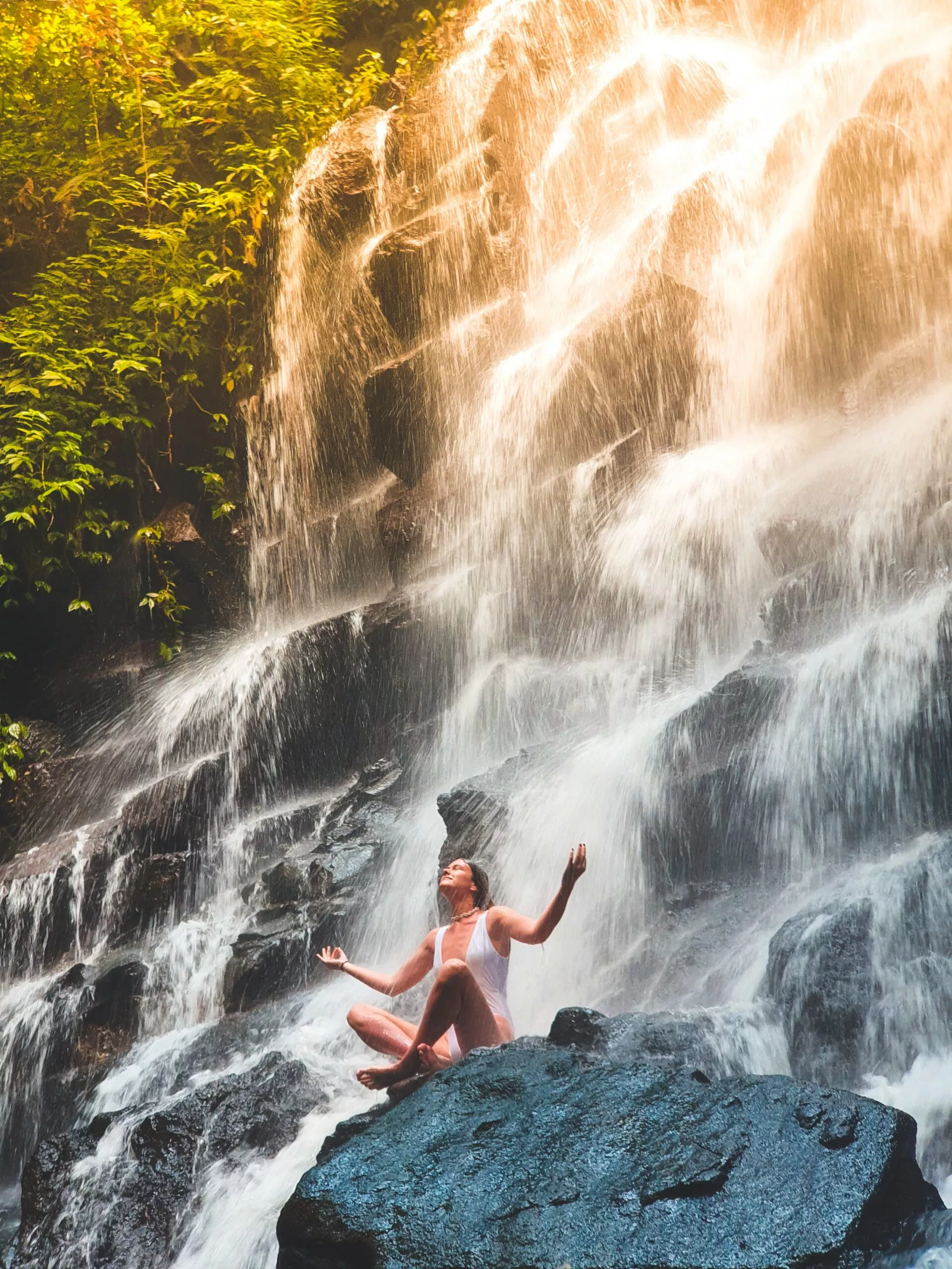 Eine Frau, die Techniken zur Stressreduktion anwendet, sitzt auf einem Felsen vor einem Wasserfall.