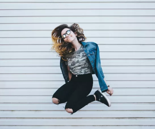 Eine Frau springt vor einer grauen Wand mit angewinkelten Beinen in die Höhe.