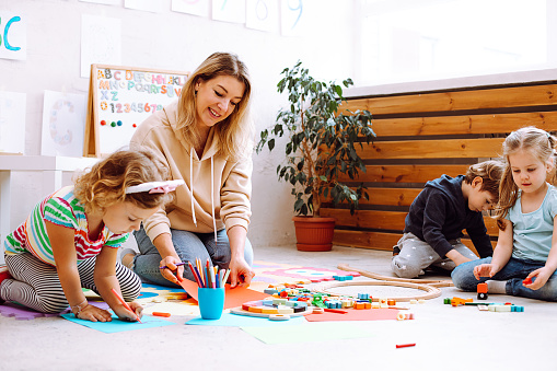 Porträt einer jungen lächelnden Lehrerin, die mit Kindern sitzt und ein Gemälde auf rotem Papier betrachtet.