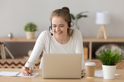 Lächelnde Frau mit Headset schreibt Notizen und studiert online auf dem Laptop