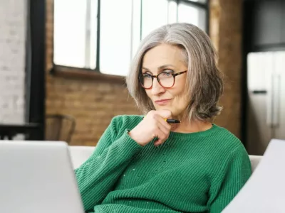 Eine ältere Frau in grüner Strickjacke arbeitet nachdenklich an einem Laptop in einer häuslichen Umgebung.