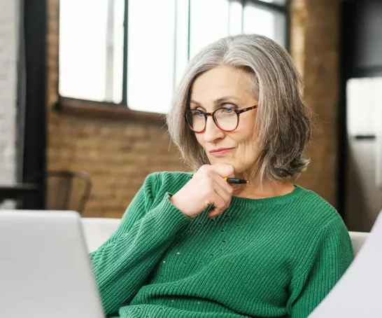 Eine ältere Frau in grüner Strickjacke arbeitet nachdenklich an einem Laptop in einer häuslichen Umgebung.