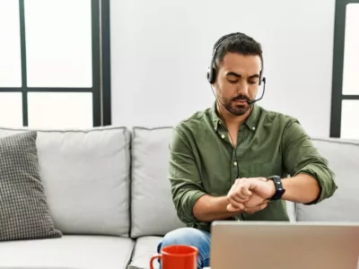 Ein Mann mit Bart und Kopfhörern schaut auf seine Uhr, während er nachdenklich an einem Laptop in seinem Wohnzimmer sitzt.
