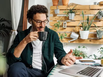 Ein Mann mit lockigem Haar und Brille sitzt konzentriert an einem Laptop in einer gemütlichen, grünen Umgebung mit Zimmerpflanzen und Holzmöbeln. Er trinkt aus einer Tasse, während er an seinem Laptop arbeitet.