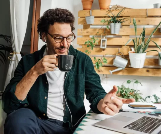 Ein Mann mit lockigem Haar und Brille sitzt konzentriert an einem Laptop in einer gemütlichen, grünen Umgebung mit Zimmerpflanzen und Holzmöbeln. Er trinkt aus einer Tasse, während er an seinem Laptop arbeitet.