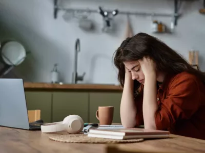 Eine Frau sitzt gestresst und erschöpft an ihrem Laptop, umgeben von Arbeitsmaterialien.