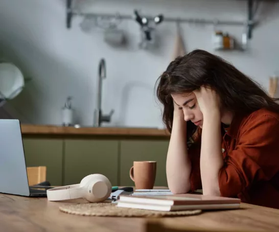 Eine Frau sitzt gestresst und erschöpft an ihrem Laptop, umgeben von Arbeitsmaterialien.