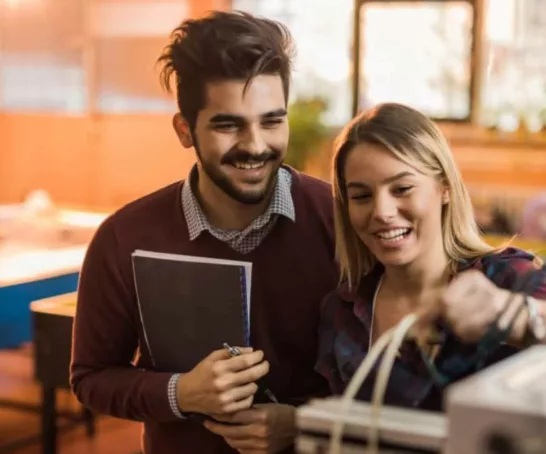 Zwei Studierende, die zusammen an einer Maschine arbeiten. Sie wirken glücklich und zufrieden bei ihrer gemeinsamen Arbeit.