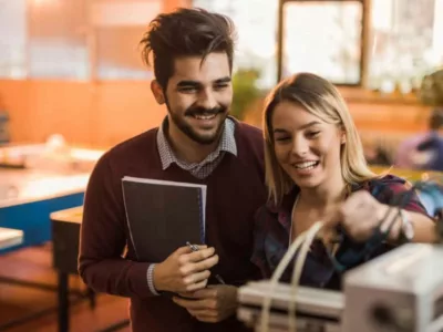 Zwei Studierende, die zusammen an einer Maschine arbeiten. Sie wirken glücklich und zufrieden bei ihrer gemeinsamen Arbeit.