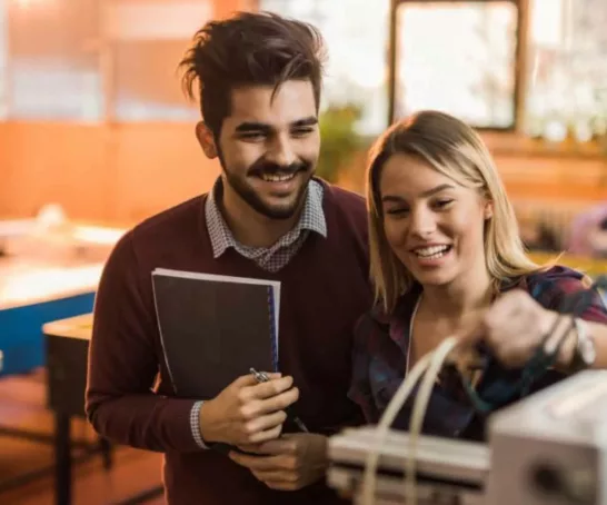 Zwei Studierende, die zusammen an einer Maschine arbeiten. Sie wirken glücklich und zufrieden bei ihrer gemeinsamen Arbeit.
