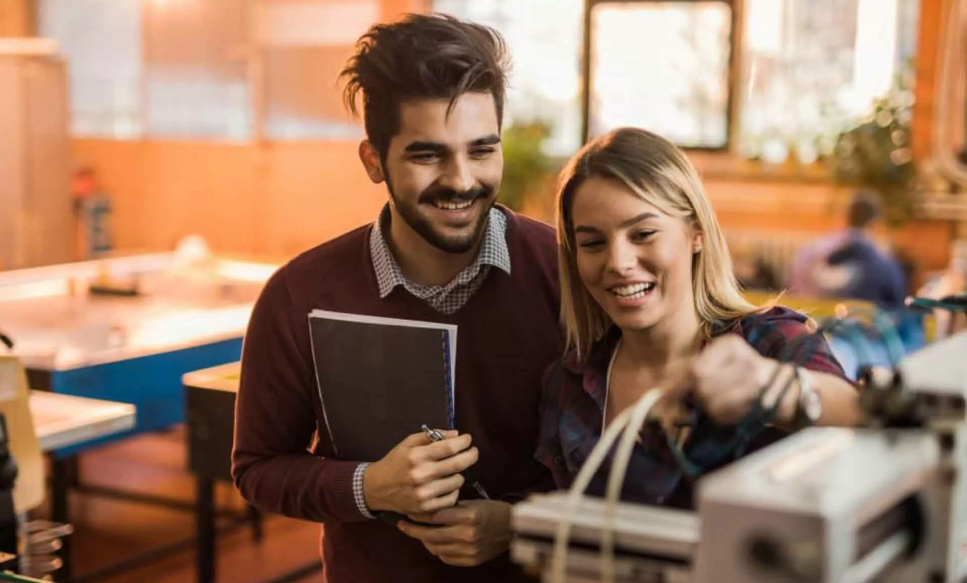 Zwei Studierende, die zusammen an einer Maschine arbeiten. Sie wirken glücklich und zufrieden bei ihrer gemeinsamen Arbeit.