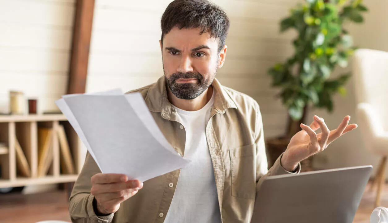 Das Bild zeigt einen Mann mit Bart, der offensichtlich frustriert ist, während er an einem Laptop in seinem Homeoffice arbeitet.