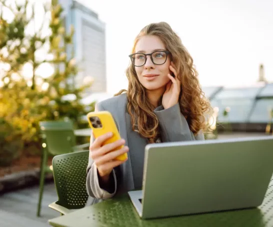 Studentin, die an einem Café sitzt. Sie trägt einen grauen Blazer und hat lockiges braunes Haar.