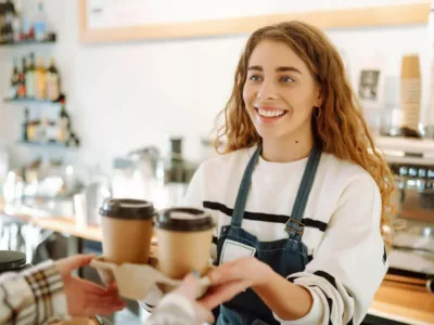 Eine Studentin arbeitet in einem Café. Sie trägt eine weiße Bluse und eine schwarze Schürze und reicht 2 Kaffeegetränke über den Tresen.