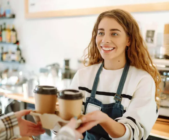 Eine Studentin arbeitet in einem Café. Sie trägt eine weiße Bluse und eine schwarze Schürze und reicht 2 Kaffeegetränke über den Tresen.