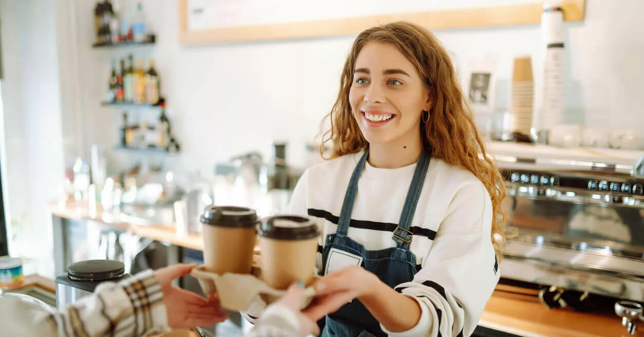 Eine Studentin arbeitet in einem Café. Sie trägt eine weiße Bluse und eine schwarze Schürze und reicht 2 Kaffeegetränke über den Tresen.