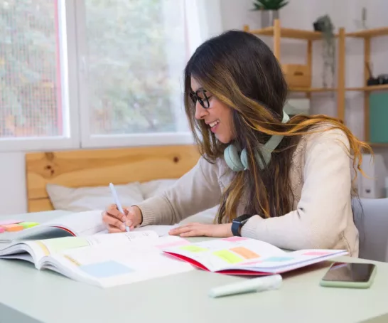 Eine Studentin mit langen dunklen Haaren sitzt an einem Schreibtisch in einem Heimbüro und lernt konzentriert, während sie eifrig Notizen macht.