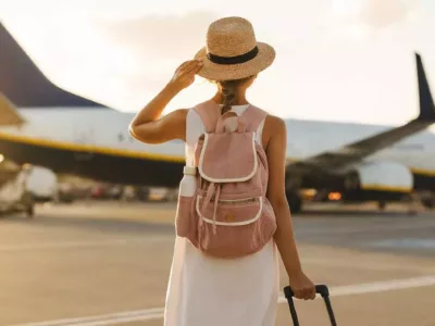 Eine Frau mit Strohhut und Rucksack steht vor einem Flugzeug auf dem Rollfeld und schaut zum Himmel auf.