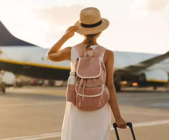 Eine Frau mit Strohhut und Rucksack steht vor einem Flugzeug auf dem Rollfeld und schaut zum Himmel auf.