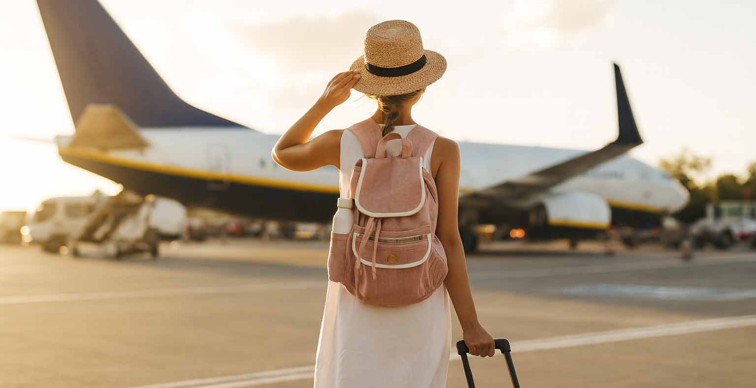 Eine Frau mit Strohhut und Rucksack steht vor einem Flugzeug auf dem Rollfeld und schaut zum Himmel auf.