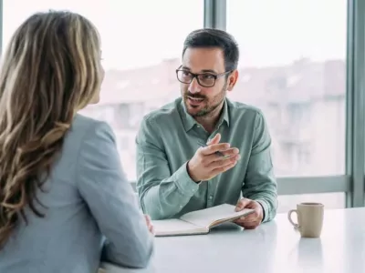 Kolleg:innen sitzen an einem Tisch und führen ein Gespräch. Der Mann mit Brille und grünem Hemd scheint etwas in einem Buch oder Dokument zu zeigen, während die Frau aufmerksam zuhört.