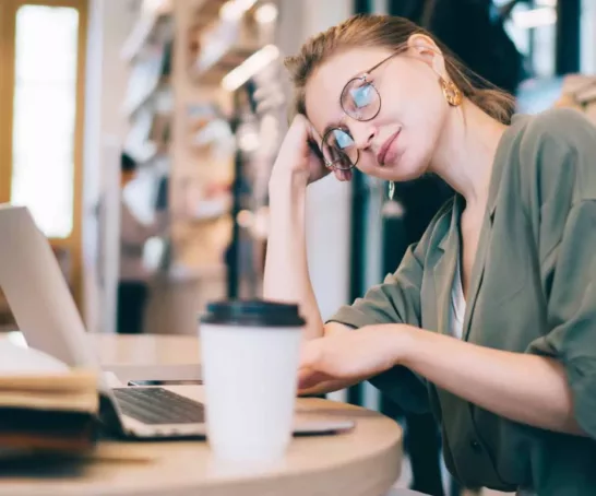 Studentin, die in einer Bibliothek oder einem Arbeitsbereich an einem Schreibtisch sitzt. Sie stützt ihren Kopf mit den Händen ab und sieht müde oder frustriert aus.