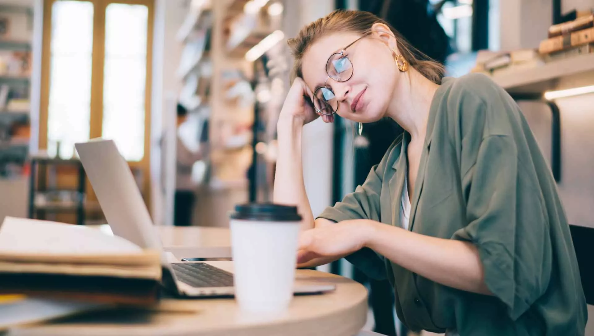 Studentin, die in einer Bibliothek oder einem Arbeitsbereich an einem Schreibtisch sitzt. Sie stützt ihren Kopf mit den Händen ab und sieht müde oder frustriert aus.