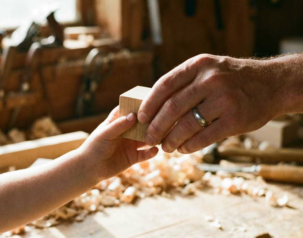 Es zeigt Hände, die einen Holzbaustein übergeben.