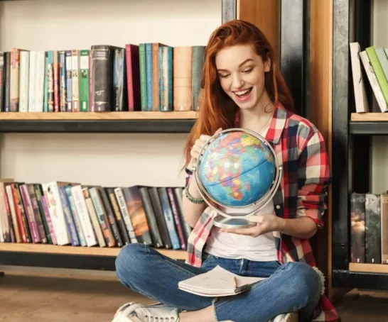 Junge Frau sitzt auf dem Boden, hält einen Globus in der Hand und im Hintergrund steht ein Bücherregal.