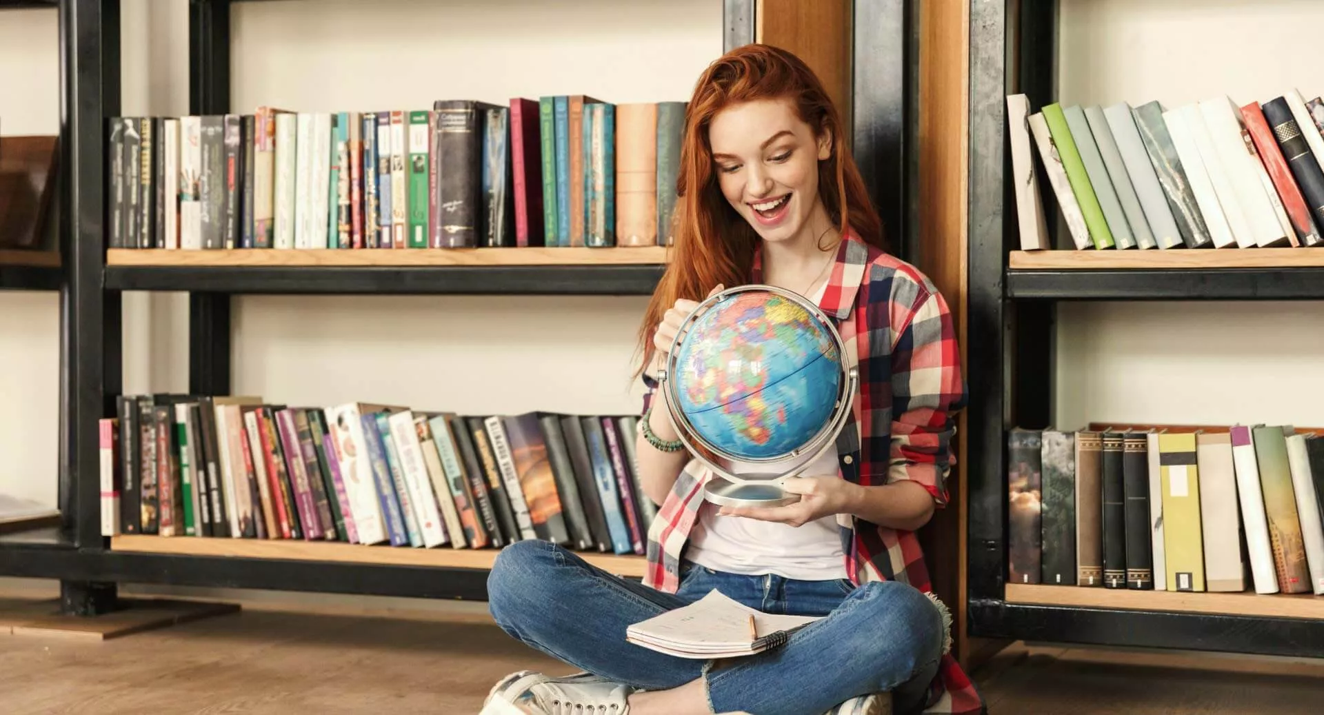 Junge Frau sitzt auf dem Boden, hält einen Globus in der Hand und im Hintergrund steht ein Bücherregal.