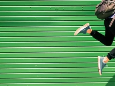 Student mit Rucksack springt hoch, im Hintergrund ist eine grüne Holzwand.