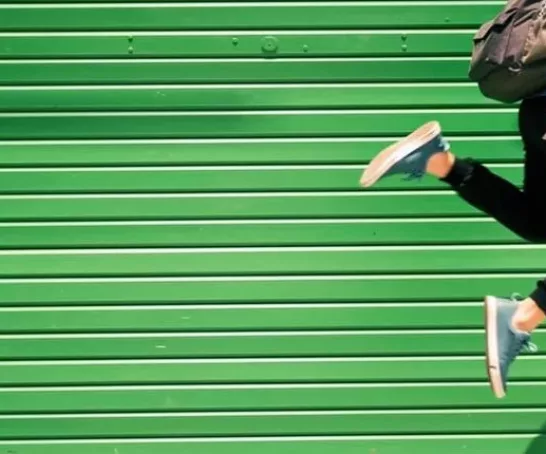 Student mit Rucksack springt hoch, im Hintergrund ist eine grüne Holzwand.