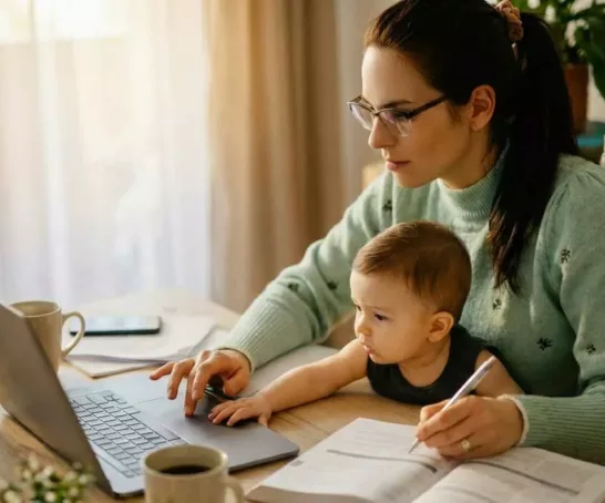 Eine Studentin mit Brille und hellgrünem Pullover sitzt an einem hölzernen Schreibtisch und arbeitet an einem Laptop, während ein kleines Kleinkind neben ihr sitzt und auf die Tastatur schaut.
