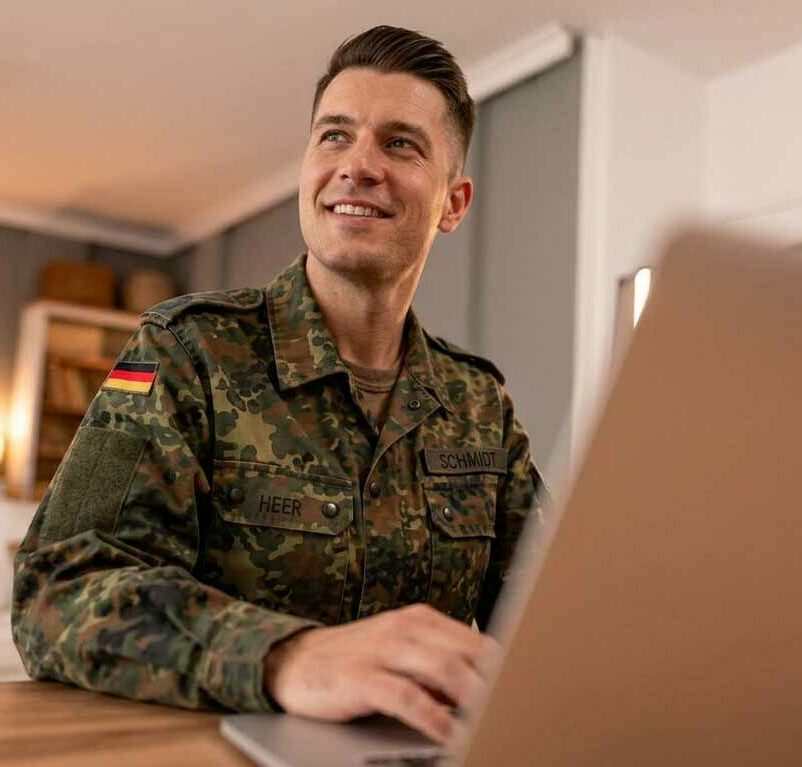 Lächelnder Soldat der Bundeswehr in Flecktarn-Uniform sitzt an einem Laptop