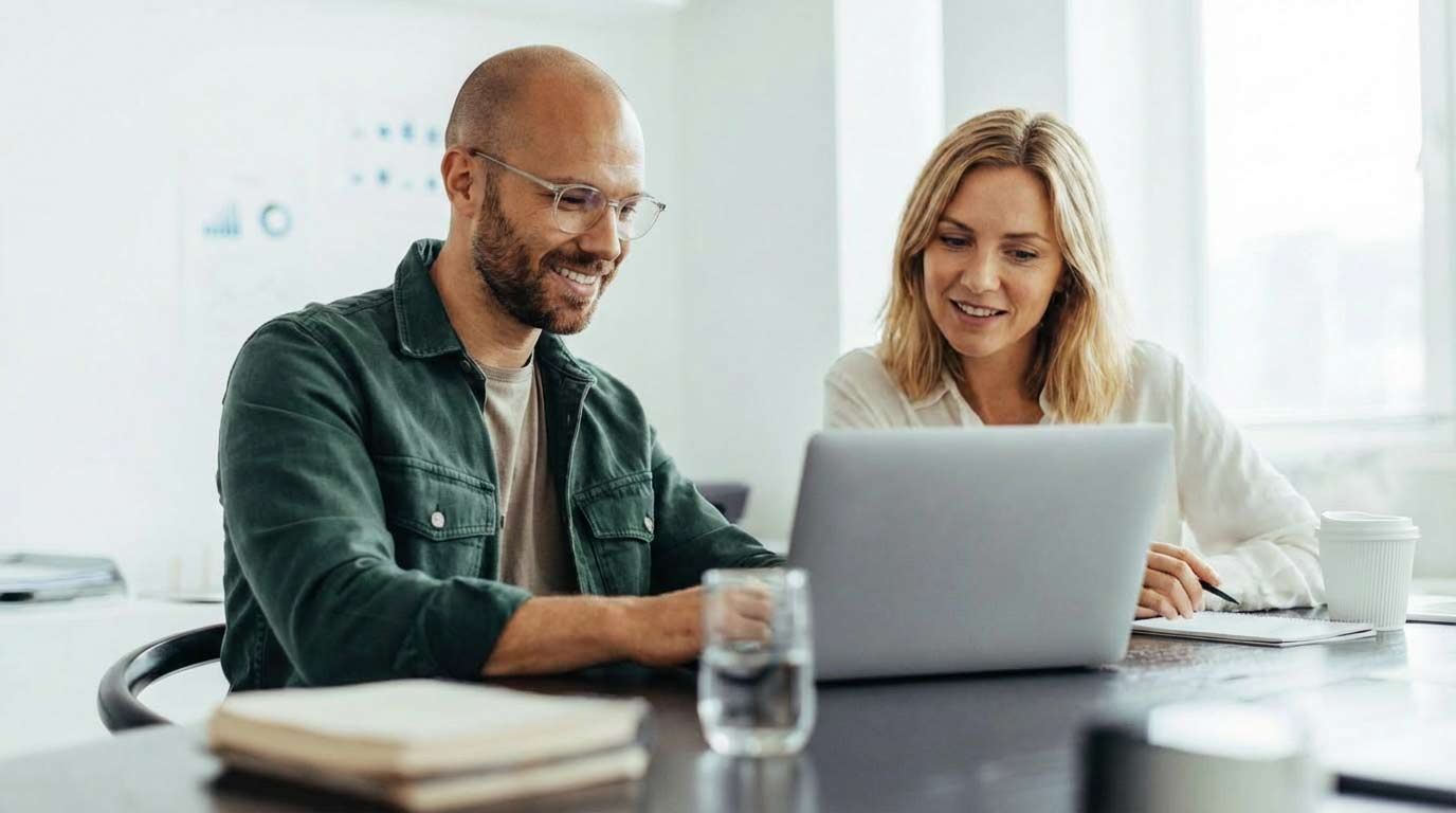 Zwei Kollegen arbeiten lächelnd zusammen an einem Laptop im modernen Büro