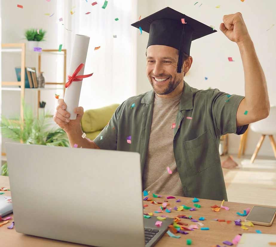 Student jubelt mit Konfetti und Talar vor seinem Laptop, mit einer Urkunde in der Hand