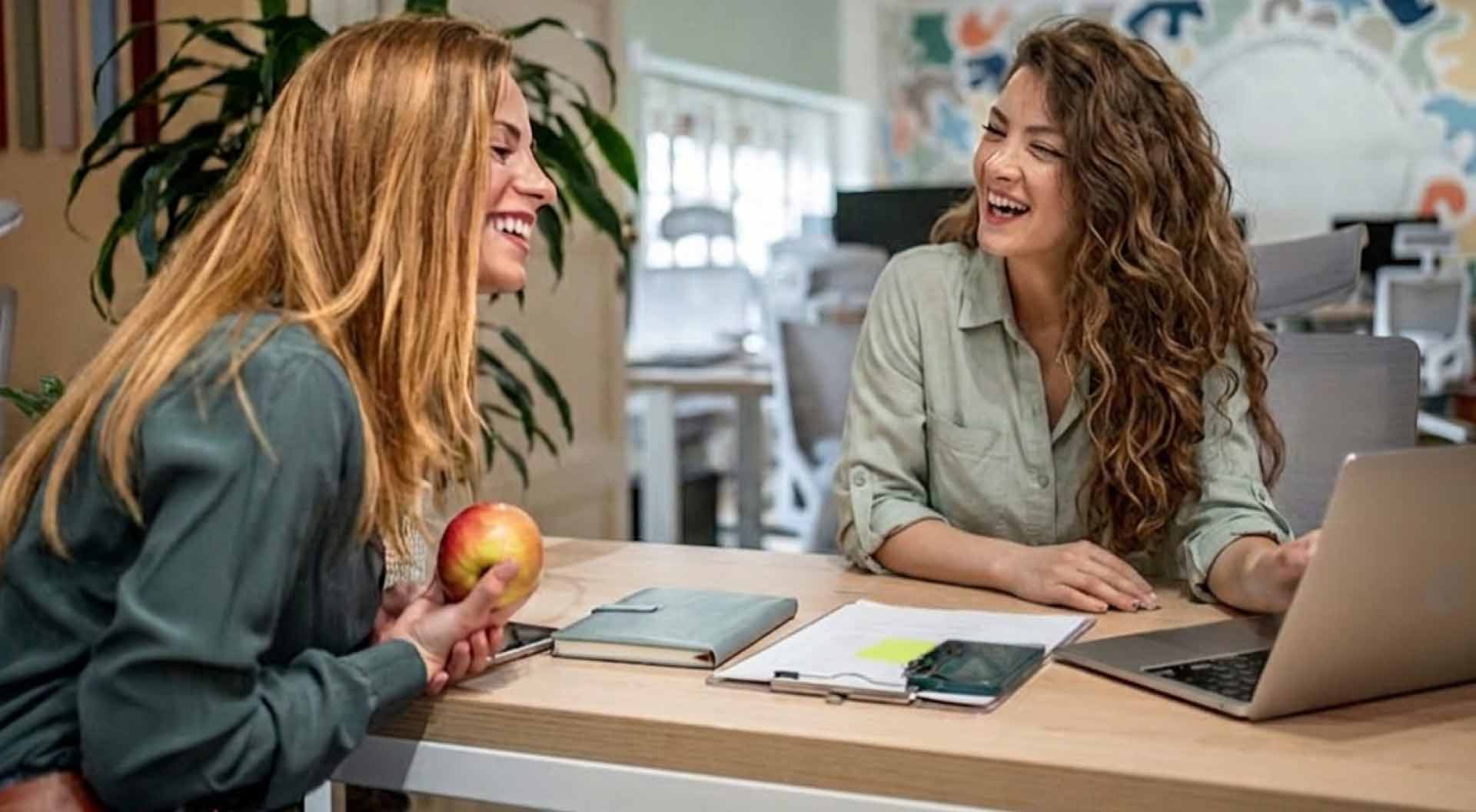 Zwei lachende Frauen mit Apfel und Notizbuch am Schreibtisch