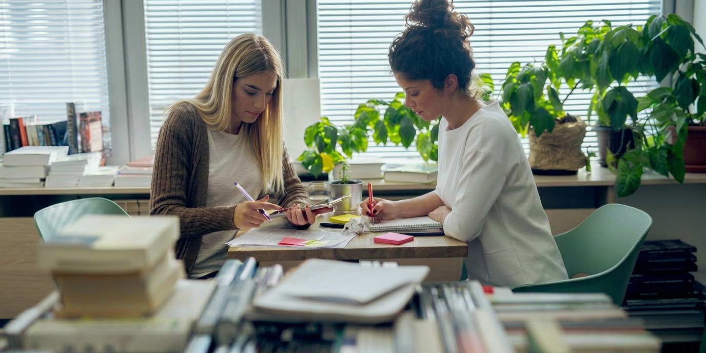 Zwei Frauen schreiben gemeinsam am Schreibtisch mit Pflanzen und Büchern.
