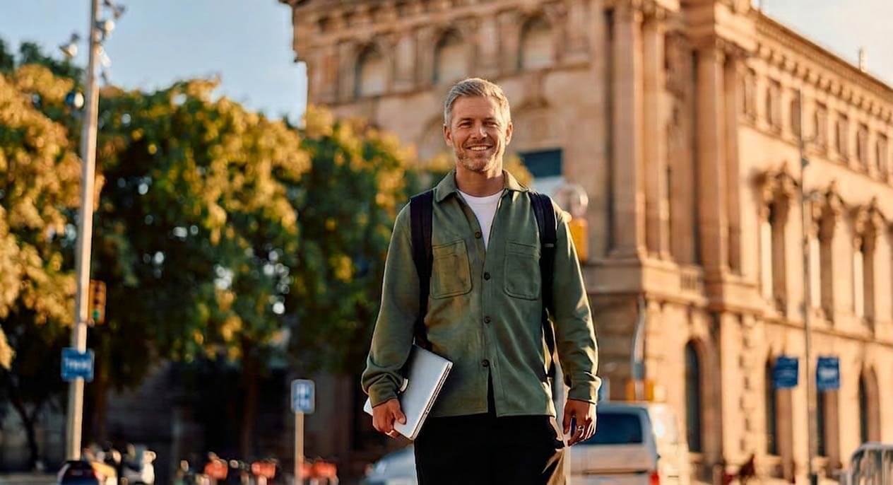 Ein lächelnder Mann mittleren Alters in olivfarbener Jacke mit Laptop steht vor historischen Gebäuden in einer sonnigen Universitätsstadt.