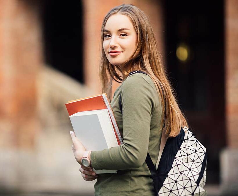Junge Frau mit langen braunen Haaren in olivgrünem Shirt hält ein rot-weiß kariertes Buch und trägt einen schwarzen Rucksack, lächelt über die Schulter vor verschwommenem Backsteingebäude.