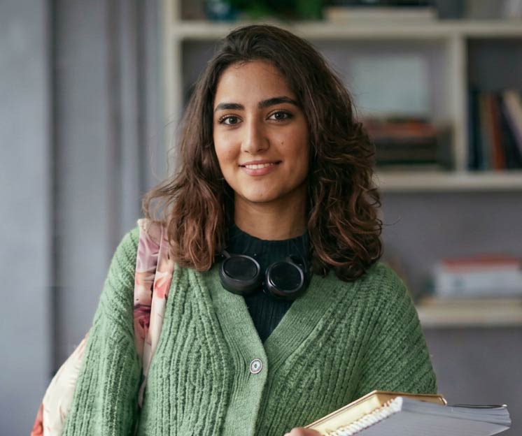 Junge Frau mit dunklen, welligen Haaren trägt einen grünen Strickjacke und schwarzes Oberteil, hält ein Notizbuch und lächelt zur Kamera