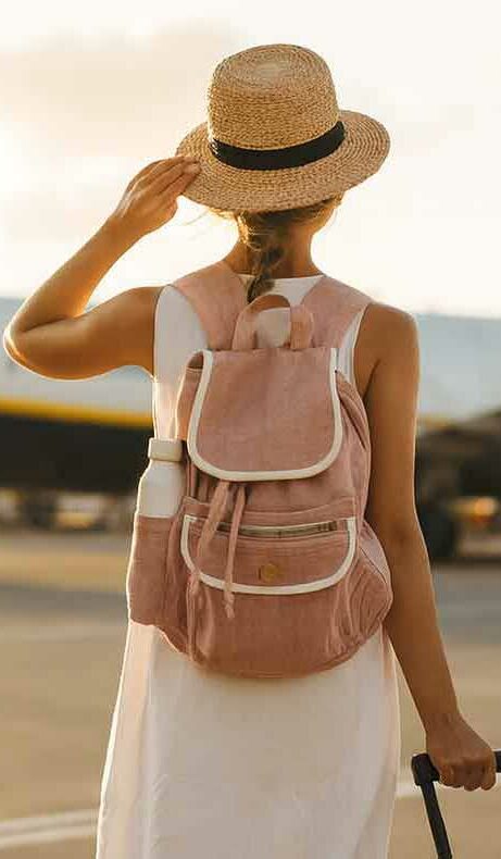 Eine Frau mit Strohhut und Rucksack steht vor einem Flugzeug auf dem Rollfeld und schaut zum Himmel auf.