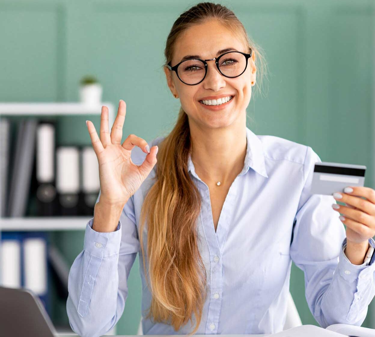 Lächelnde Geschäftsfrau im Büro hält Kreditkarte mit OK-Geste.