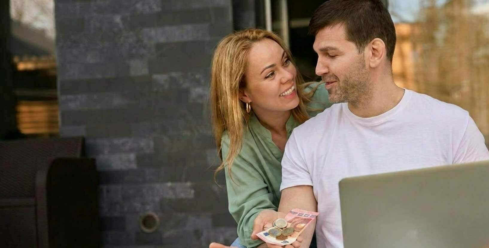 Junges Paar überprüft Finanzen auf Laptop während es Bargeld in der Hand hält.