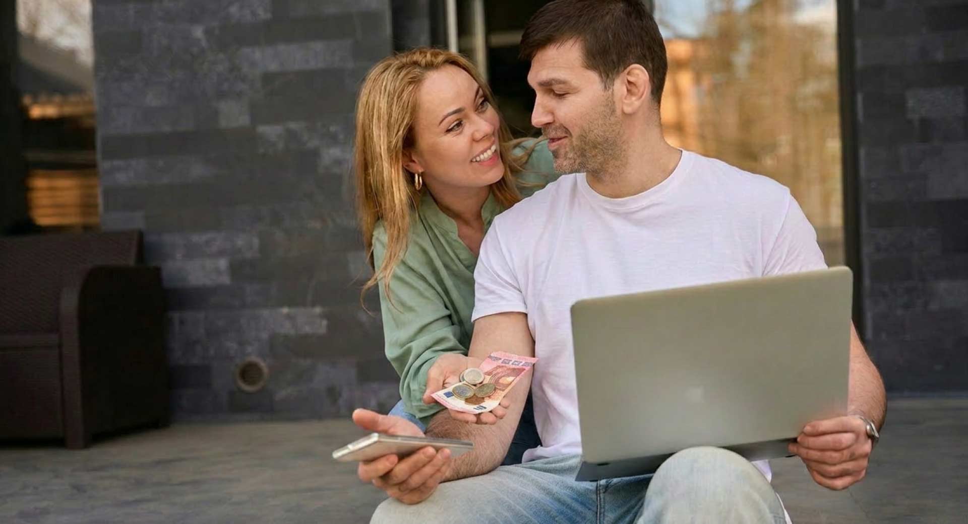 Junges Paar überprüft Finanzen auf Laptop während es Bargeld in der Hand hält.