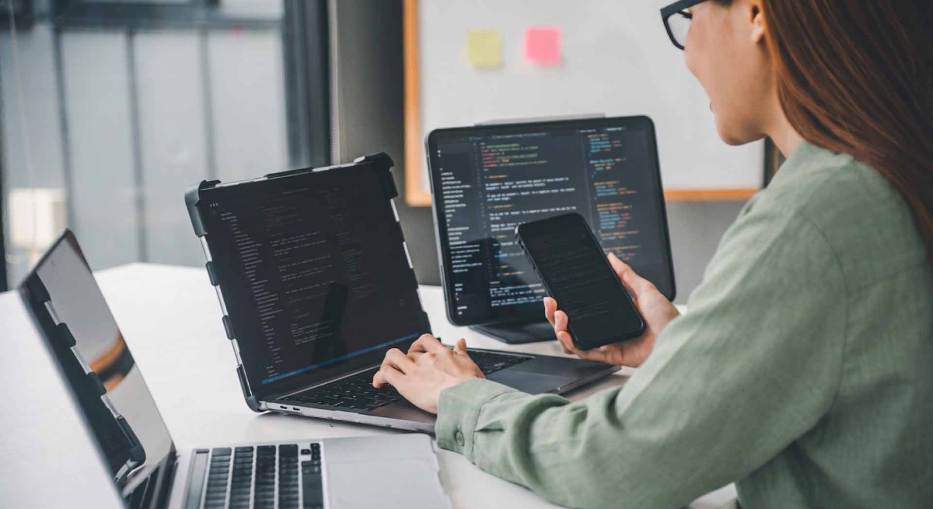 Eine Informatikstudentin arbeitet an ihrem Schreibtisch mit mehreren Geräten: Laptop, Tablet und Smartphone mit Programmiercode auf den Bildschirmen.