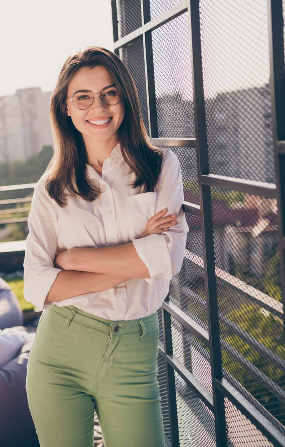 Junge Frau mit Brille und braunen Haaren steht lächelnd auf einem Balkon. Sie trägt ein weißes Hemd und grüne Hose. Stadtlandschaft im Hintergrund.