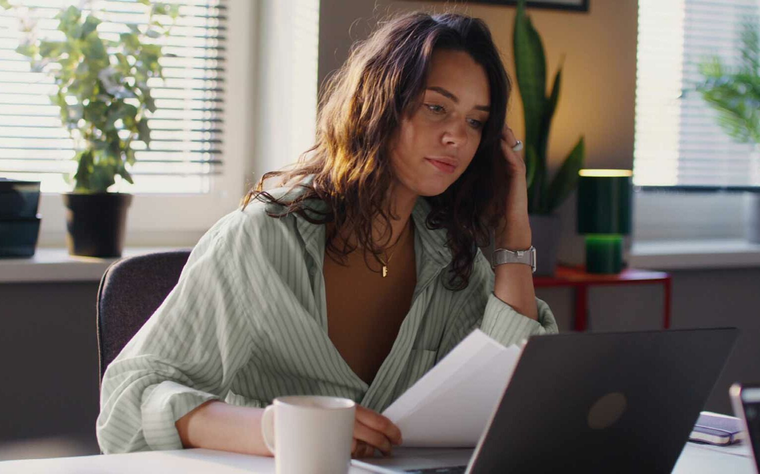 Eine Frau mit Handy und Kaffeetasse sitzt am Schreibtisch vor einem Laptop.