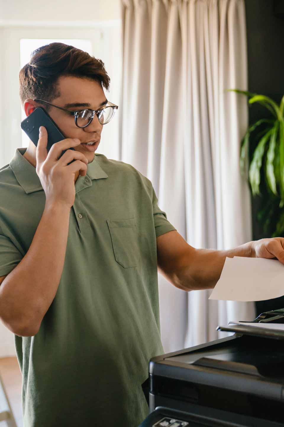 Ein junger Mann mit Brille und grünem Poloshirt hält ein Smartphone ans Ohr und schaut auf ein Dokument.